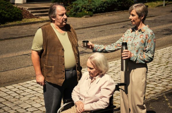eine ältere Frau sitzt im Rollstuhl. Hinter ihr ein dicklicher Mann in Tshirt und Weste sowie eine schlanke mittelalte grauhaarige Frau, die dem Mann aus ihrer Thermoskanne etwas zu Trinken anbietet